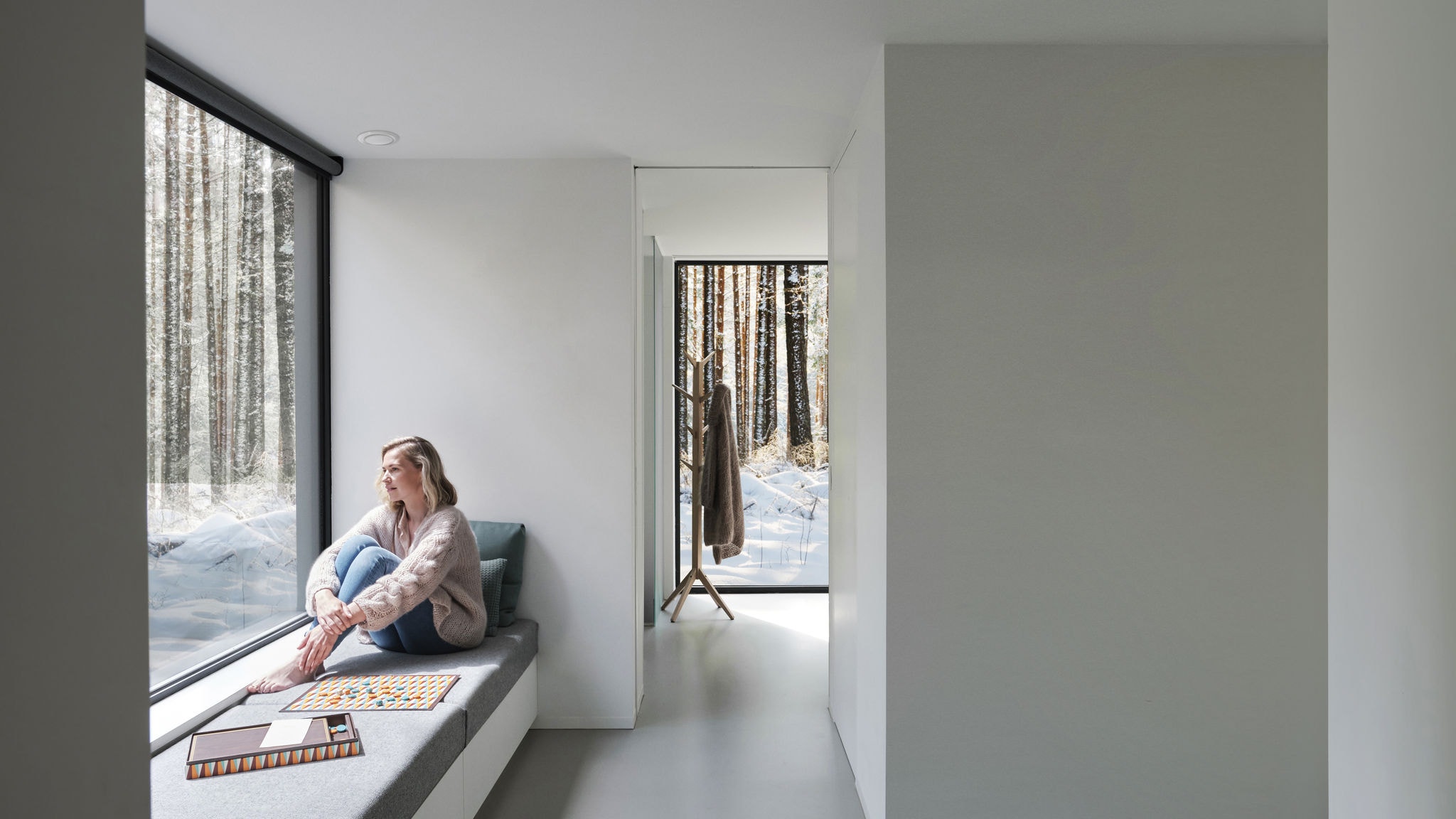 Широк кадар со жена што седи пред прозорецот боса и гледа кон дрвата и снегот.