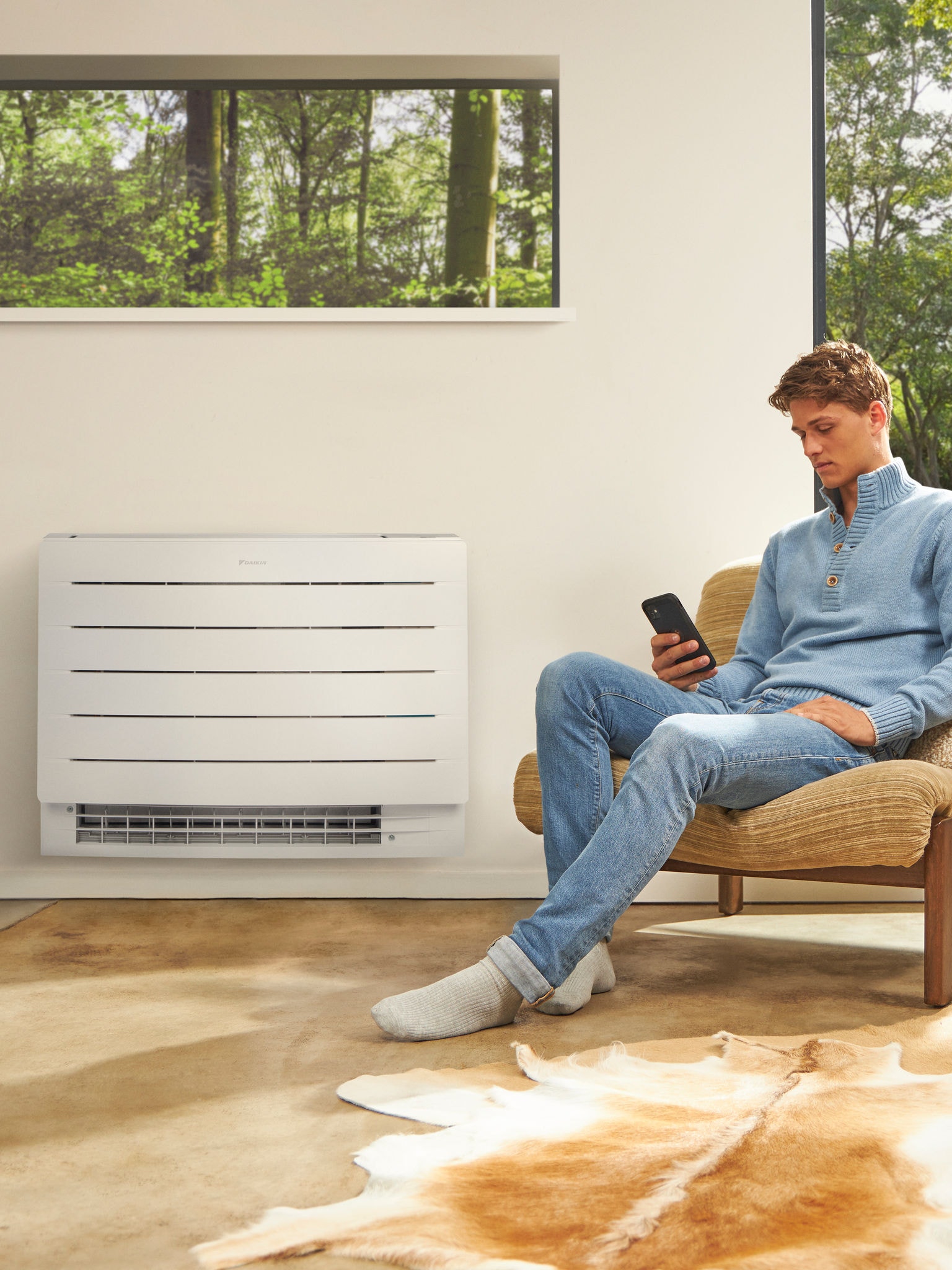 Човек седи во светла просторија и гледа во својот телефон, а зад него на ѕидот се наоѓа топлинска пумпа