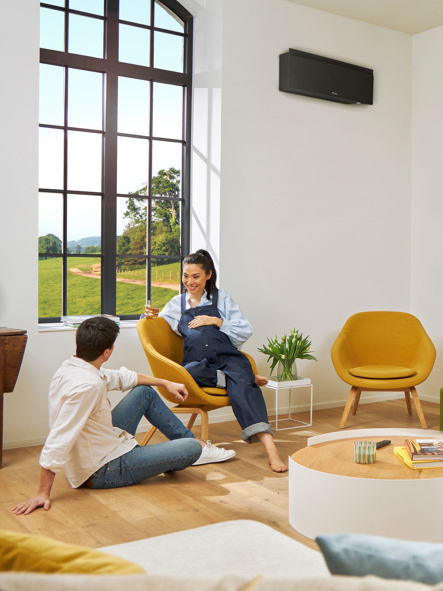 Маж и жена седат пред голем прозор во осветлена просторија со црна високоѕидна топлинска пумпа на ѕидот зад нив