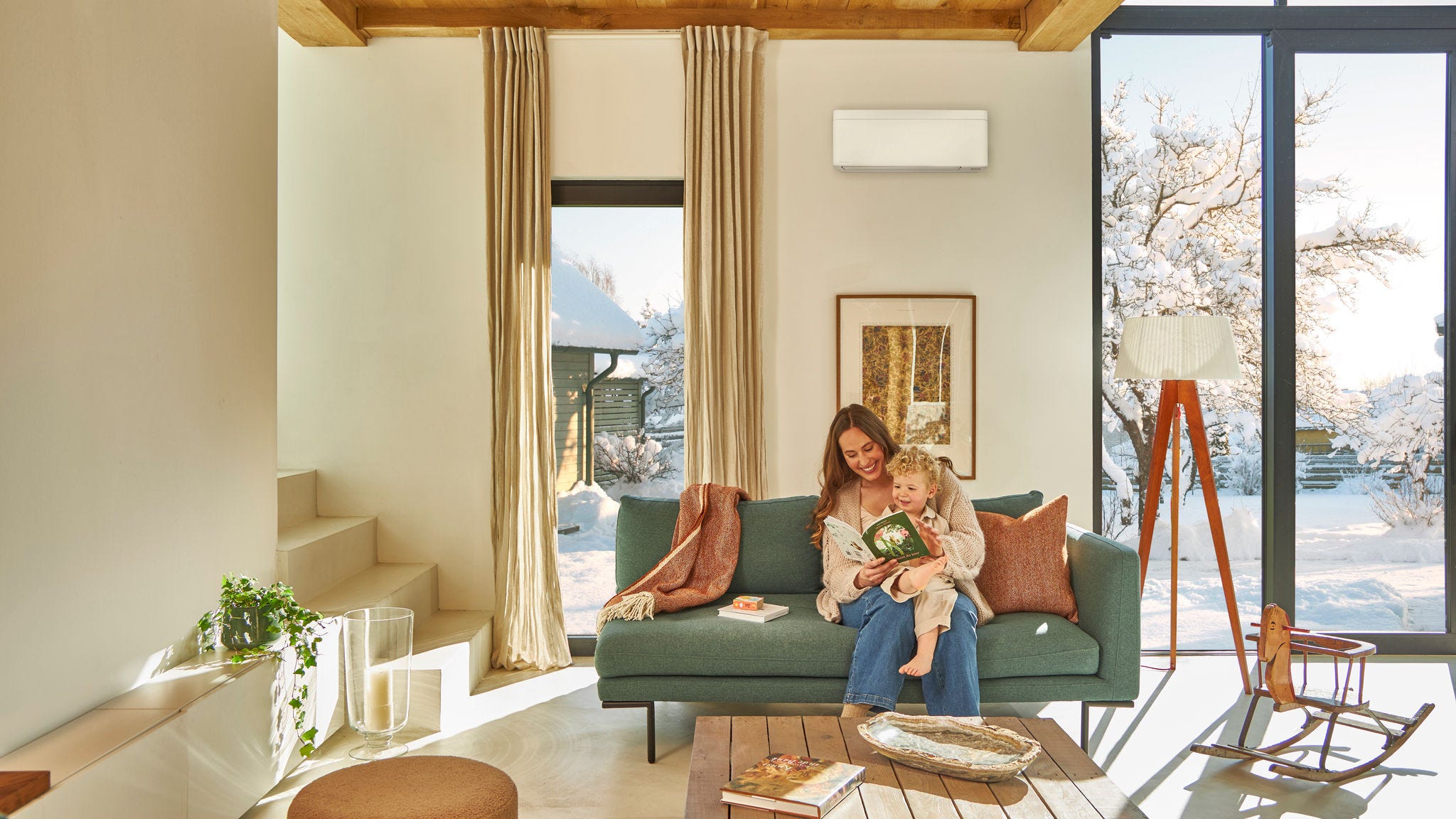 Мајка седи на каучот под високоѕидна топлинска пумпа воздух-воздух и читаат детска книга со своето малечко на возраст од 12-36 месеци на преносен компјутер, уживајќи во топлината додека надвор паѓа снег